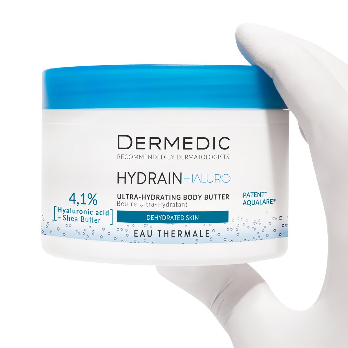 A gloved hand holds a white and blue jar of Dermedic Hydrain Ultra-Hydrating Body Butter, labeled for dehydrated skin with 4.1% hyaluronic acid and shea butter.