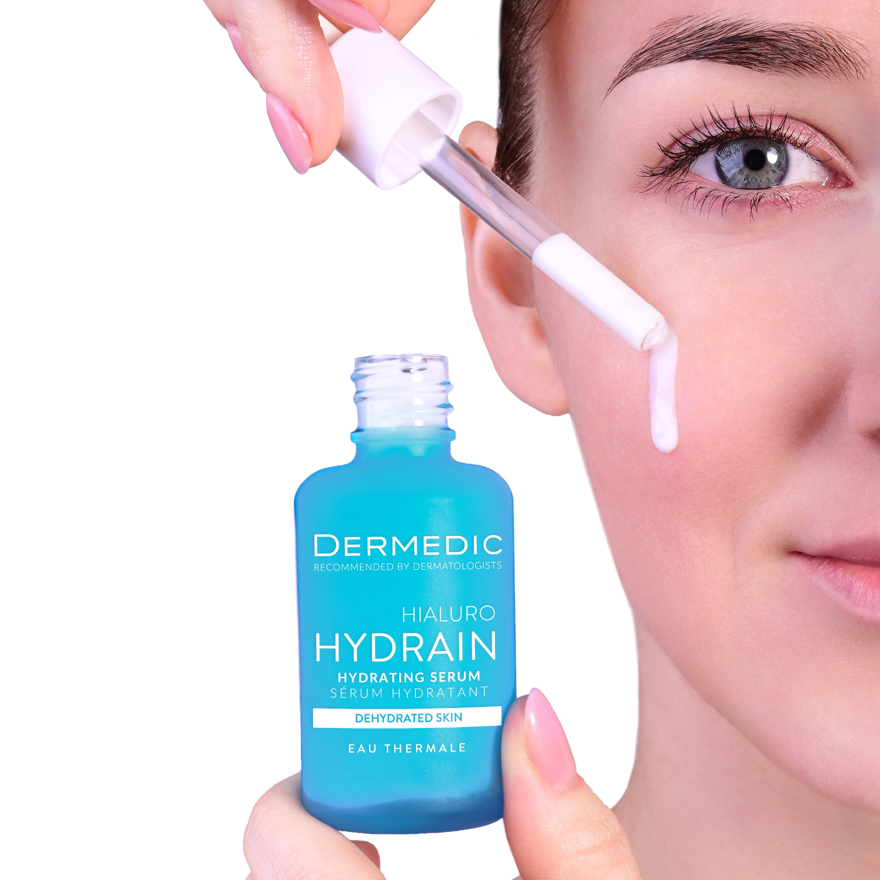 A person applies Dermedic Hydrain hydrating serum to their cheek using a dropper, with a close-up of their face and the blue serum bottle in focus.