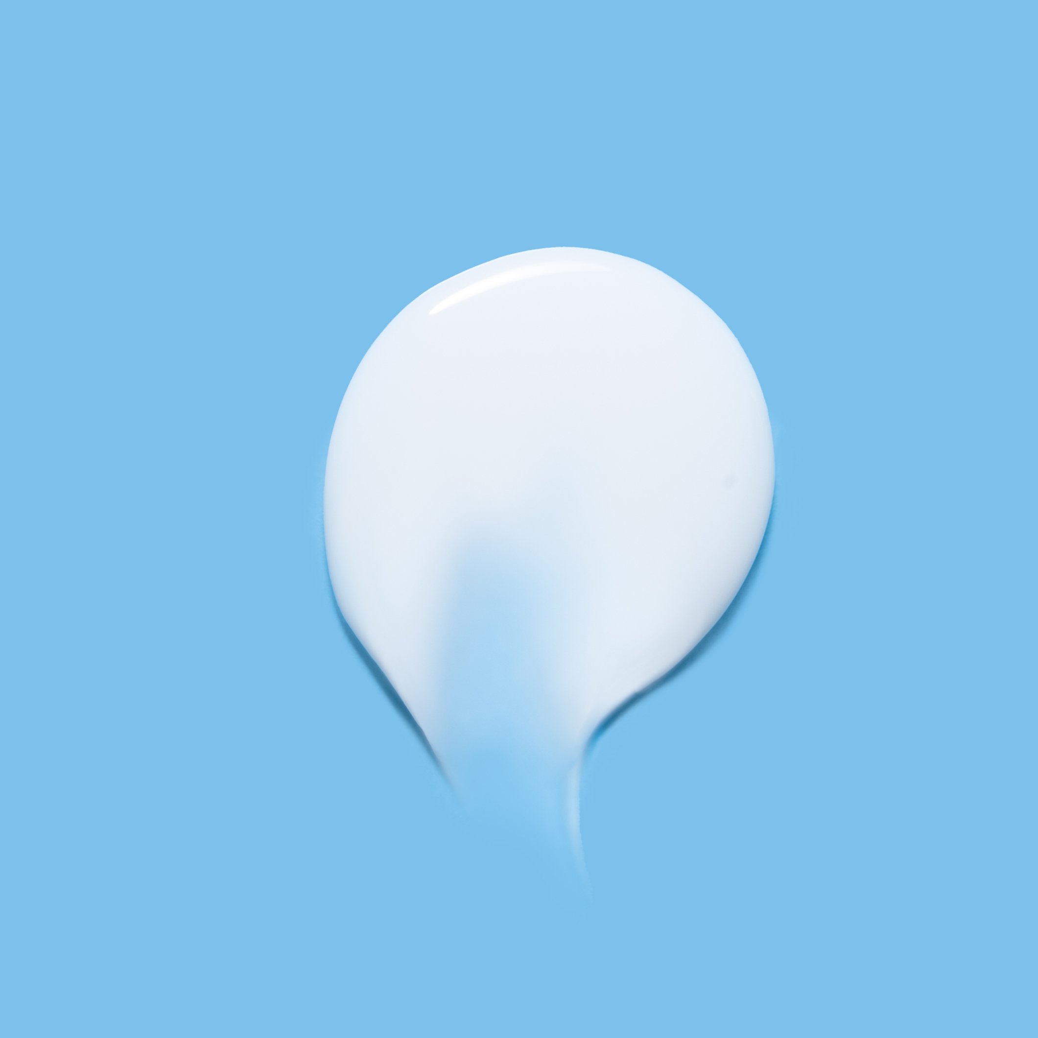 A smear of white cream or lotion is spread on a bright blue background, creating a smooth, curved shape with soft shadows.