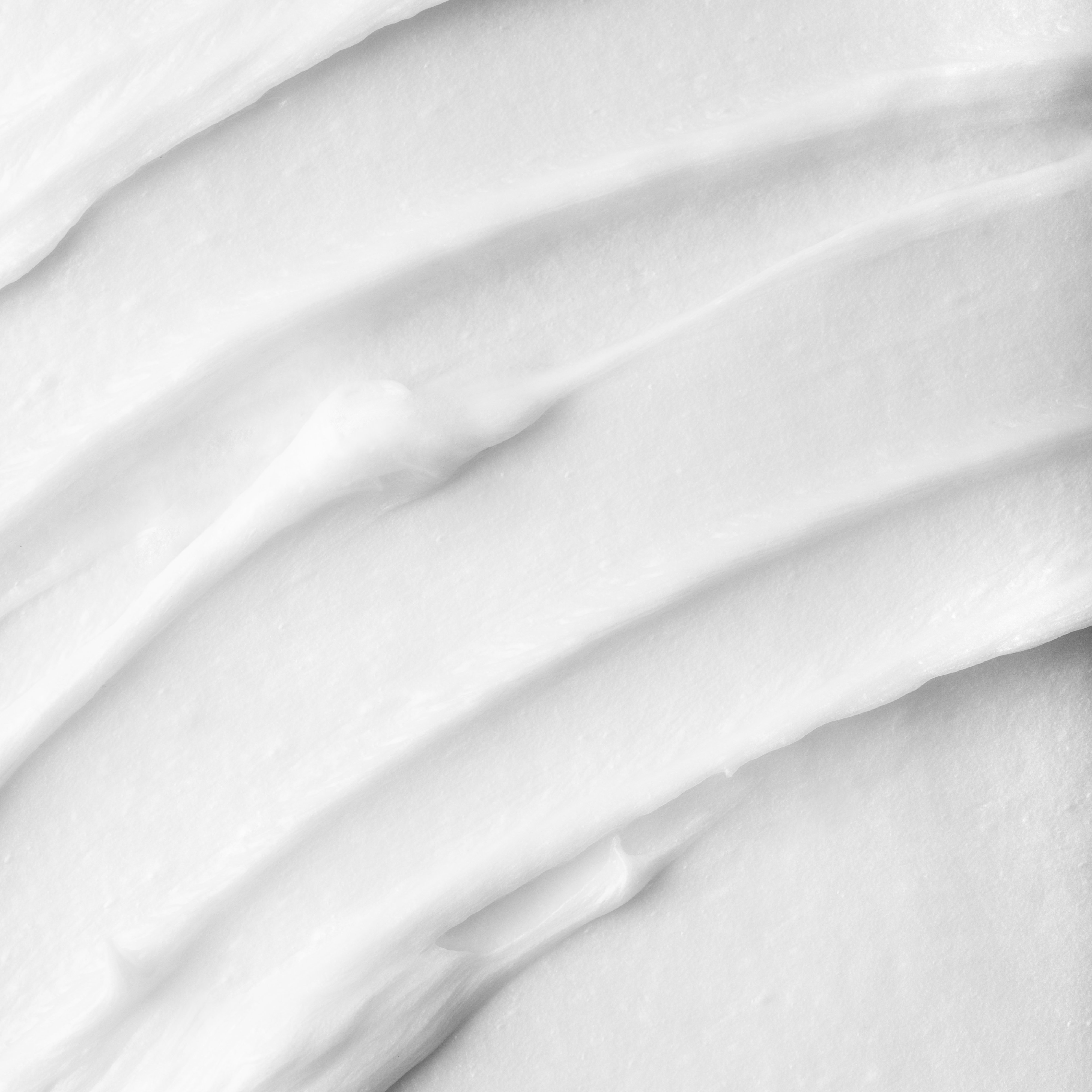Close-up of white creamy substance with smooth, textured, and slightly wavy surface, resembling lotion, cream, or whipped product spread evenly.