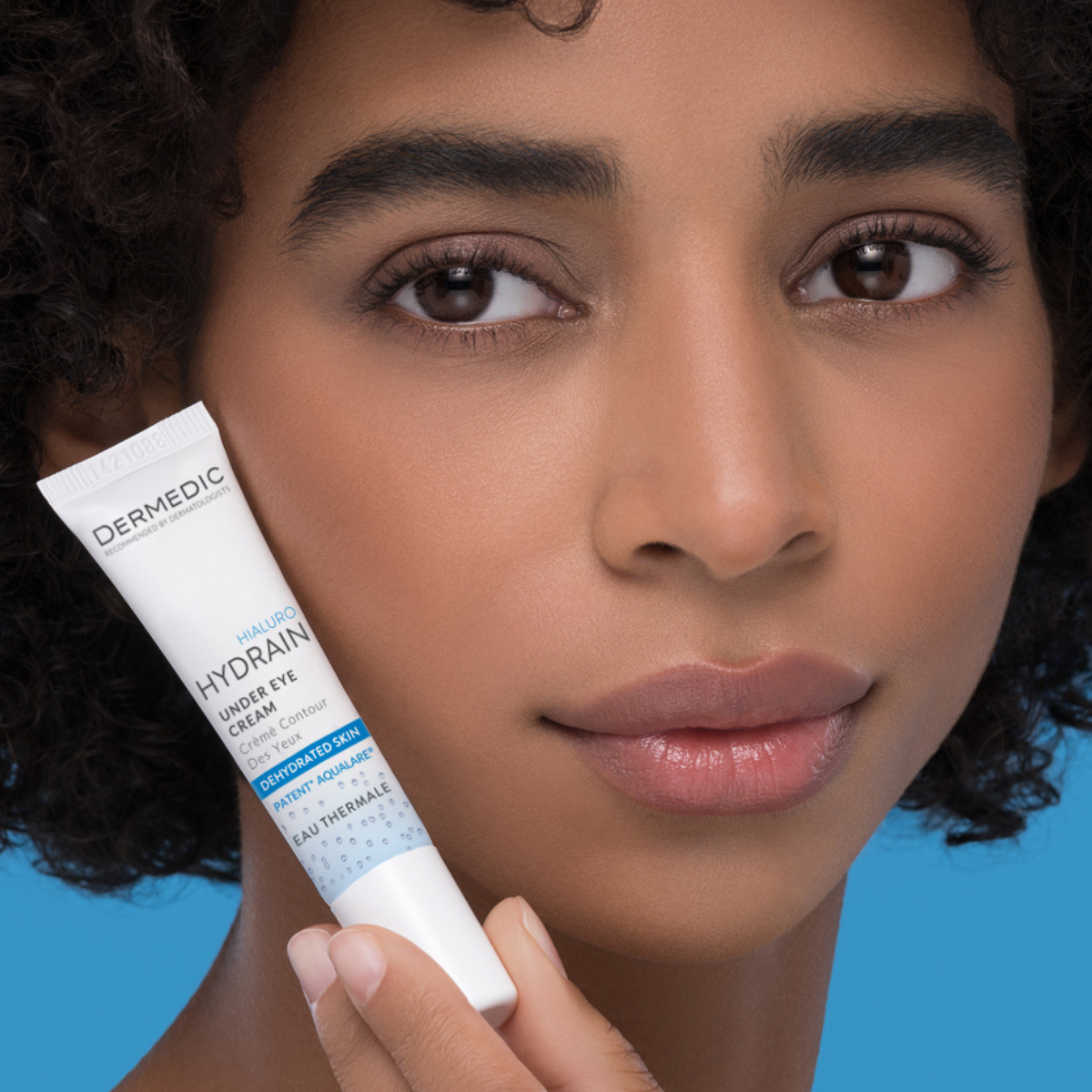 A woman with curly hair holds a tube of Dermedic Hydrain under eye cream close to her face, with a smooth complexion and a blue background.